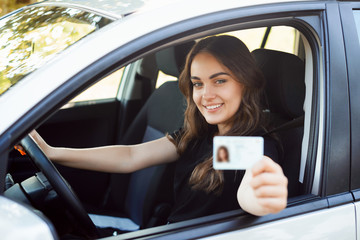 Happy student driver sitting in the modern silver car and showing driving car license to the camera...