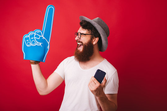 Portrait Of Amazed Bearded Man Celebrating Success Holding Smartphone And Foam Finger