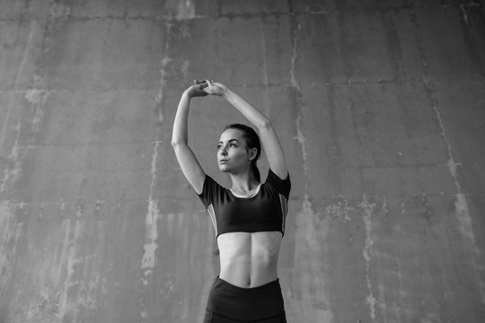 Sports woman on urban gray background. Fitness model working out outdoor. Beautiful slim brunette girl doing stretching warmup exercise. Black and white.
