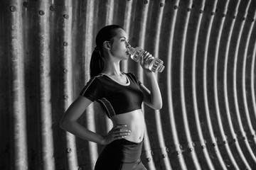 Fototapeta na wymiar Beautiful slim brunette woman drinking water from bottle after running. Fitness young model working out outdoor on tunnel urban gray background. Black and white.