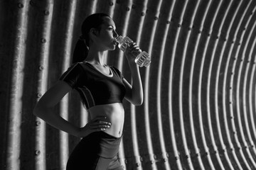 Fototapeta na wymiar Beautiful slim brunette woman drinking water from bottle after running. Fitness young model working out outdoor on tunnel urban gray background. Black and white.