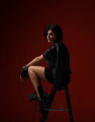 Thoughtful woman in black tight dress, leather jacket and big brutal boots sitting on a stepladder and looks down on red