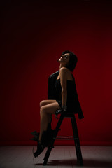 Young sensual woman in black tight dress, leather jacket and big brutal boots resting on a stepladder on dark red