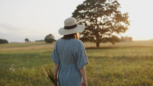 Pregnant woman walking in meadow during sunset. Pretty mother to be dressed in linen dress and straw hat with flower bouquet in the field by oak tree takes pleasure and turns to camera and smiles