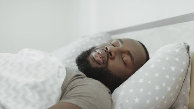 Black man sleeping in bed in morning. Young adult covering head with pillow.