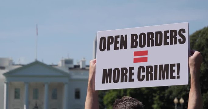A man holds an Open Borders Equals More Crime protest sign in front of the White House on a sunny summer day.	