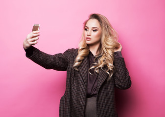 Blond girl taking photo makes self portrait on smartphone over pink background