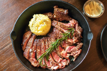 Cast iron plate with sliced grilled rib-eye cooking a medium-rare steak with mashed potato and sauce in saucer over table.