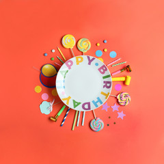 Different birthday party items and empty plate on coral background, flat lay. Space for text