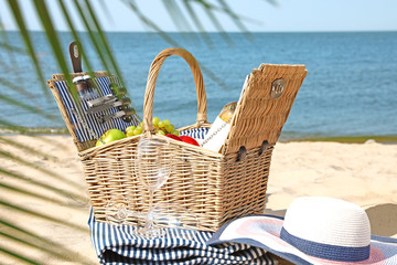 Wicker picnic basket with products, blanket and hat on sunny beach