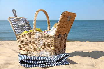 Wicker picnic basket with products and blanket on sunny beach. Space for text