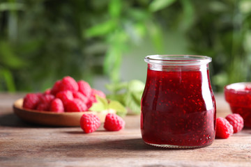 Glass jar of sweet jam with ripe raspberries on wooden table. Space for text