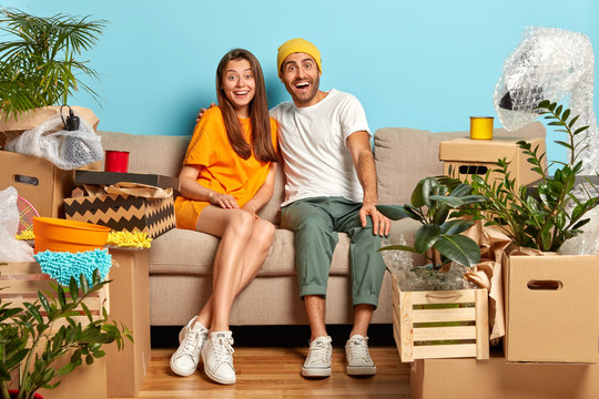 Glad Husband And Wife Look With Happiness, Hug While Sit On Sofa In Living Room, Move To New Home, Cardboard Boxes Around, Get Settled In Flat. People, Family, Moving, Repair And Real Estate Concept