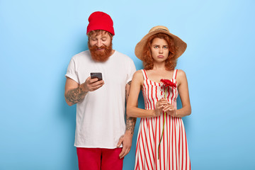 Obraz premium Unhappy redhead woman gets bored while boyfriend uses cell phone, addicted from modern technologies. Young couple have date, isolated on blue background. Bearded man with phone ignores girlfriend