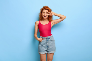 Fototapeta premium Positive red haired young woman makes peace gesture, feels lucky, has fun, keeps one hand in pocket of denim shorts, grins at camera, expresses joyful attitude, models over blue wall. Body language