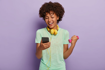 Half length shot of happy dark skinned female fitness instructor checks song in playlist, listens music in headphones, exercises with dumbbell, works on biceps has positive look, wears t shirt