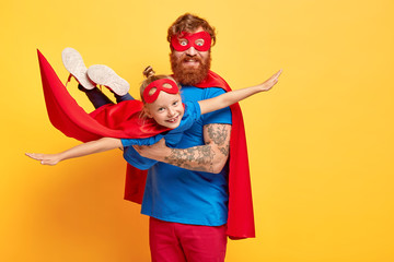 Small kid plays superman, being on fathers hands, pretends flying, spreads arms sideways, wears red mask and cloak, enjoys spare time with dad, isolated on yellow wall. Daddy hero holds daughter - obrazy, fototapety, plakaty
