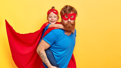 Serious bearded ginger father superhero carries small female kid on back, do good things together,...