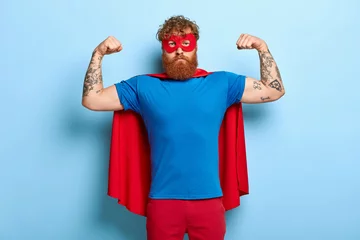 Foto op Canvas Successful hero wears red mask and cape, raises arms, shows biceps, demonstrates courage and strength, looks serious and confident, poses against blue wall. Real superhero ready to help you. © Wayhome Studio
