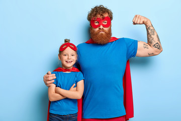 Success, power and courage concept. Superhero father shows muscles, embraces small child, dressed...