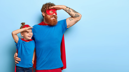 Cheerful daddy and small female child pretend being superheroes, wear blue t shirt, red masks and...