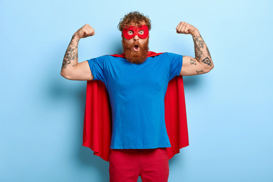 Confident red head male superhero wears mask and red cape, raises arms, shows muscles, represents power and courage, ready to defend people, has shocked expression, isolated on blue background