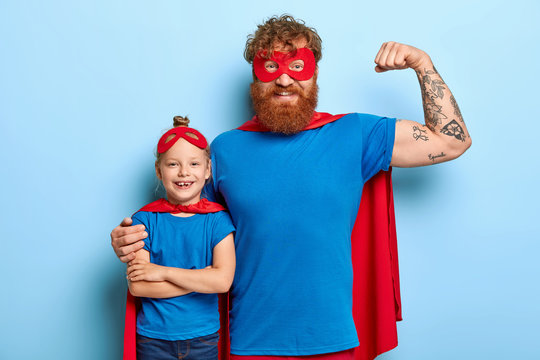 Positive optimistic daddy raises tattooed arm, shows muscles, cuddles little daughter, wear superhero suits, pretend being powerful and brave, rest together at home during spare time. Family concept