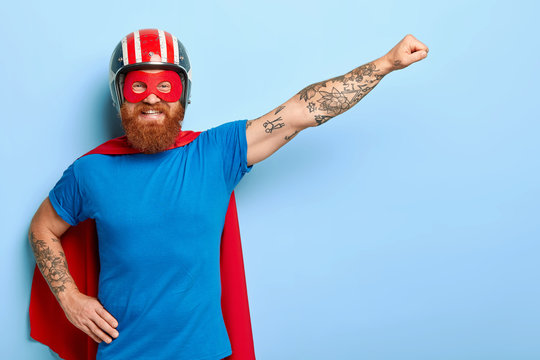 Positive bearded man has cheerful expression, keeps arm outstretched, clenches fist, ready for flight, dressed in helmet blue t shirt and cape imagines being superman uses special power to help people