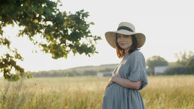 Pregnant woman dressed in linen dress and hat standing by oak tree in meadow outdoors during sunset. Beautiful mother to be relax stroking pregnant belly smiles into camera in country field