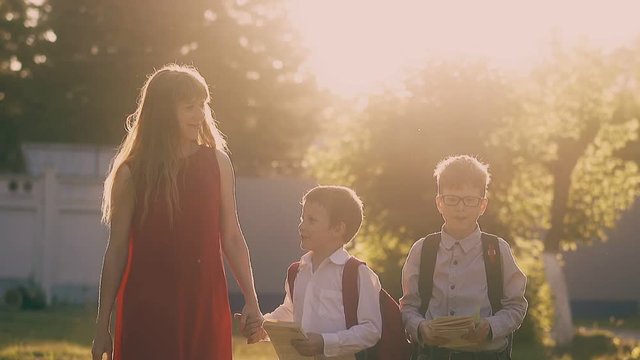 long haired lady in red dress walks holding children with school bags hands lit by bright sun slow motion
