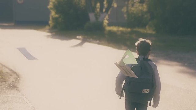 running boy with black schoolbag jumps up on grey asphalt road and throws copybooks after exam slow motion backside view