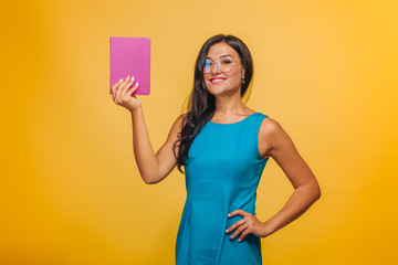 Girl with glasses on yellow background in blue dress shows pink book