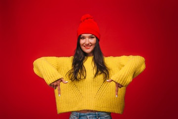 Girl in yellow sweater and hat on red background showing down