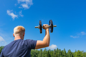 Fototapeta na wymiar Caucasian blond man stands with his back and holds a dumbbell in his right hand. The arm is raised high, the muscles are tense. Concept of sports in the open air, amateur sports. Place for text.