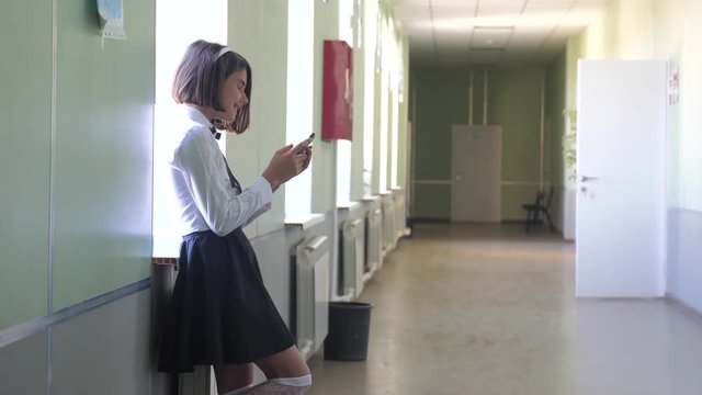schoolgirl education with smartphones at school concept. Young girl teen and lifestyle using smartphone standing by the window in the corridor of the school. correspondent in the messenger, write a