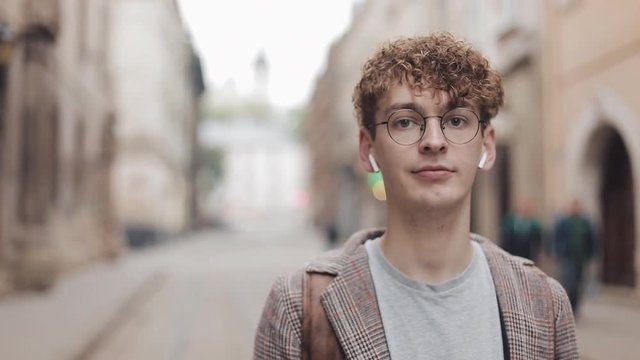 Portrait of young hipster guy with backpack standing on the city street and looking into the camera. He listens to music in wireless headphones. Tourist, travel, student concept. Urban lifestyle.