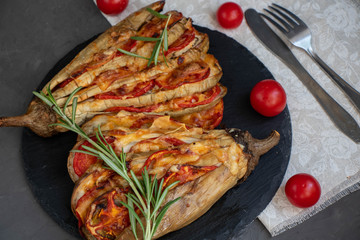 Dietary fan of eggplant white filled with tomatoes and cheese. Nearby are cherry tomatoes, rosemary branches. It is worth soy sauce and metal appliances. On a dark background.