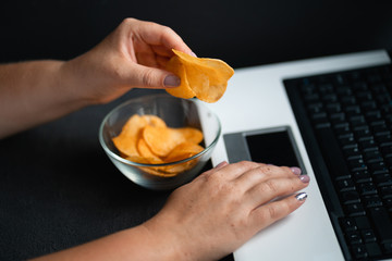 Unhealthy snack at workplace. Hands of woman working at computer and taking chips from the bowl. Bad habits, junk food, high calorie eating, weight gain and lifestyle concept