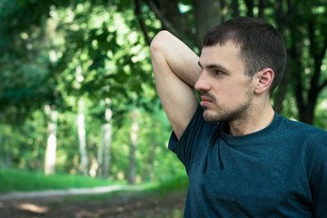 Portrait of a pensive young man in a dark green T-shirt, serious emotions, in the background a park or a forest, trees and mustaches everywhere, a hand raised up and bent at the elbow, a deep look