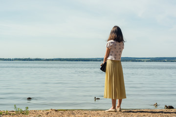 pond beach. lonely girl in a yellow and a white blouse stands on the beach and looks into the...