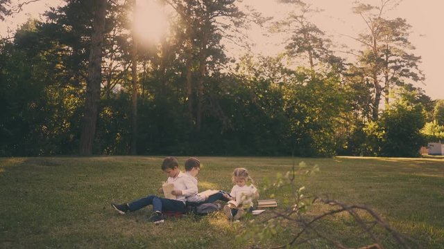 junior schoolchildren sit on green meadow during school break against green trees hiding setting sun