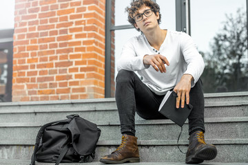 Pensive man sitting on steps of modern building and holding notepad in hands.