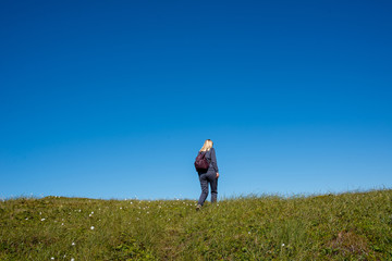 Naklejka na ściany i meble A girl with backpack outdoors rises up. Green grass and blue sky. Leisure activity, hiking. Enjoy the moment, relax. Wanderlust. Adventure, freedom, lifestyle. Explore North Norway. Summer Scandinavia