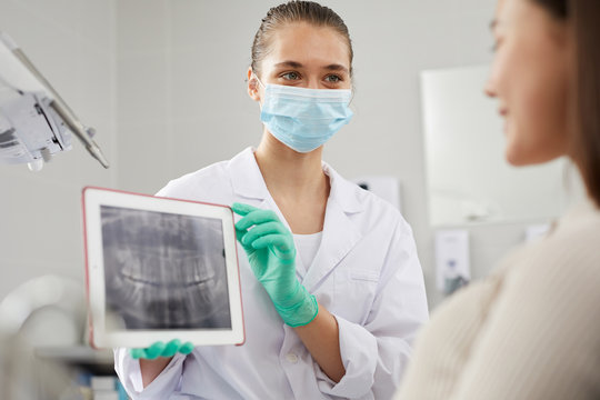 Portrait of young female dentist showing teeth x-ray image to patient during consultation in clinic, copy space