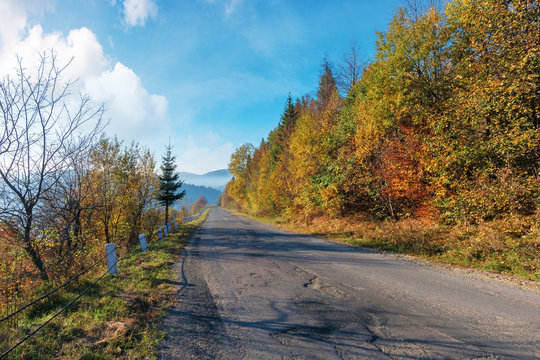 old cracked asphalt road in mountains. straight path along the forest on hill. trees in fall colors. distant ridge in haze. sunny autumn weather with fluffy clouds on the bright blue sky