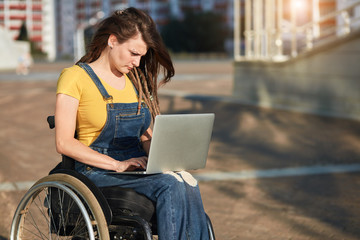 stylish disabled girl in casual clothes concentrated on working with laptop in the street, close up side view photo, copy space, blurred background.