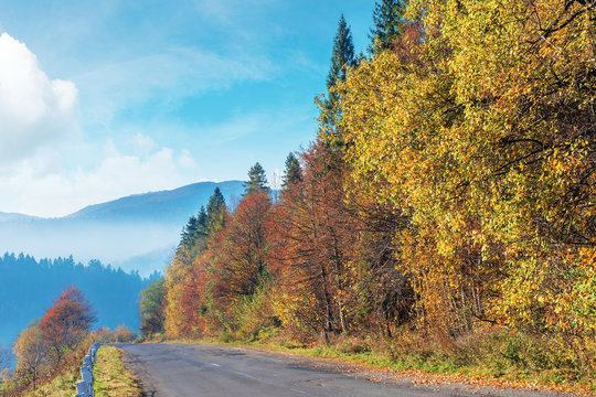 old cracked asphalt road in mountains. straight path along the forest on hill. trees in fall colors. distant ridge in haze. sunny autumn weather with fluffy clouds on the bright blue sky