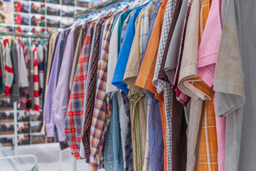 Close up view of row of colourful fabric and scotch pattern shirts  and T-shirts hang on aluminium hanger clothes rack in retail fashion store or second hand outlet shop.