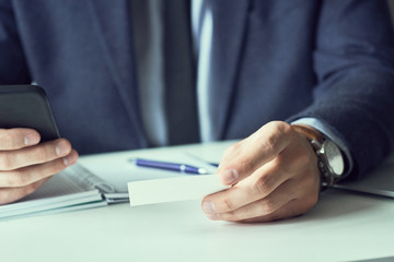 Fototapeta premium Mid section of businessman in suit holding blank business card and dialing numbers on mobile phone.