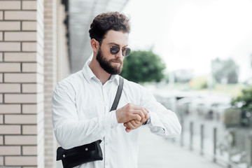 A bearded, serious, stylish man looks at the watch and walking on the streets of the city near the modern office center